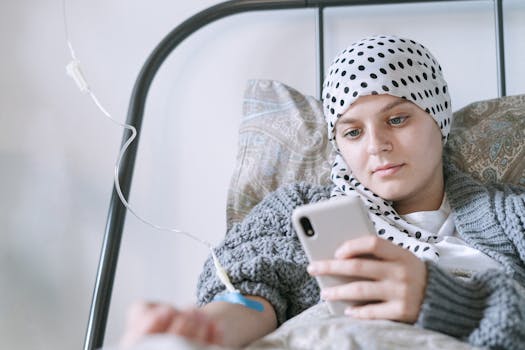 A young woman undergoing chemotherapy checks her phone in a hospital room, showcasing strength and hope.