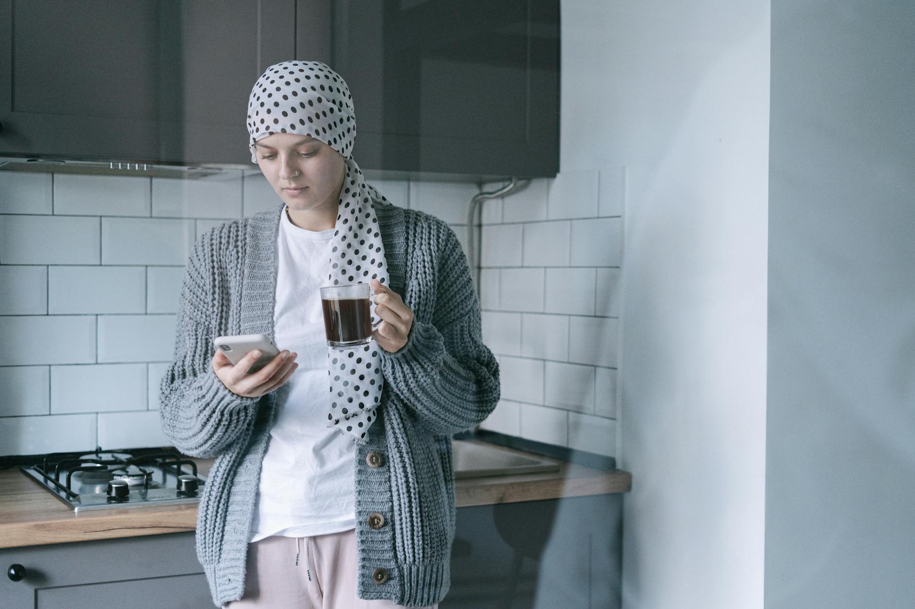A woman wearing a headscarf and sweater holds a coffee cup and phone in a modern kitchen, symbolizing strength and recovery.
