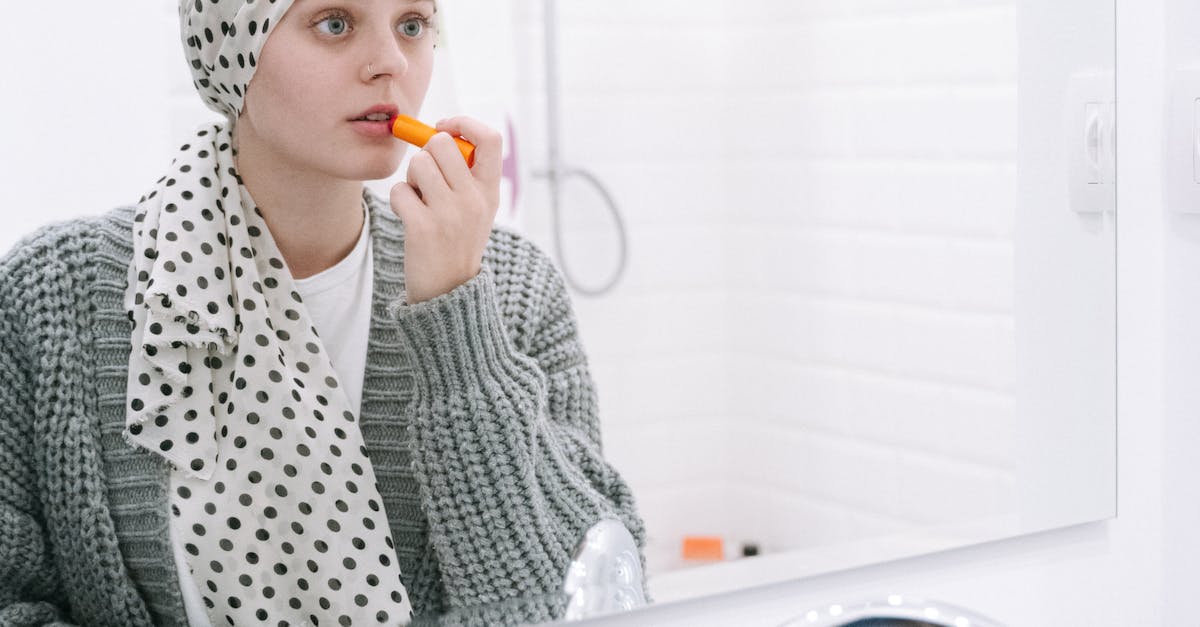Photo by Ivan S A woman wearing a headscarf applies lipstick while looking in the bathroom mirror.