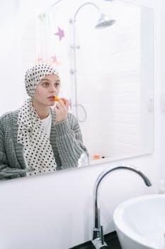 A woman wearing a headscarf applies lipstick while looking in the bathroom mirror.