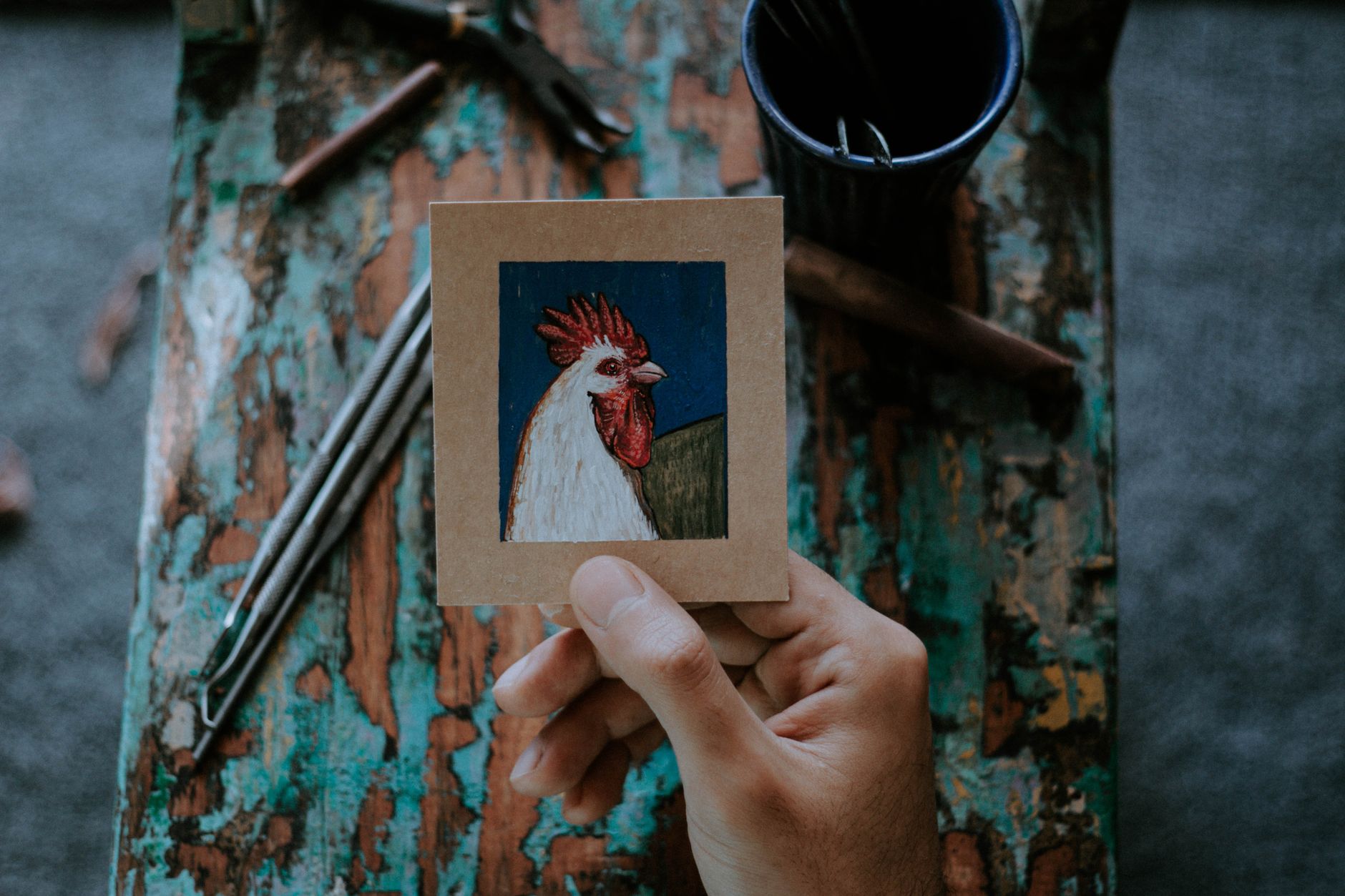 Close-up of a hand holding a rooster painting against a rustic wooden background, showcasing artistic detail.