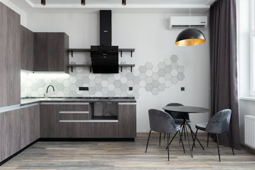 Creative design of kitchen with cupboards and cabinets under hood against table with chairs in daylight at home