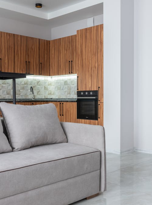 Interior of contemporary spacious light room with kitchen counter with modern oven near wooden cabinets and comfortable sofa at home