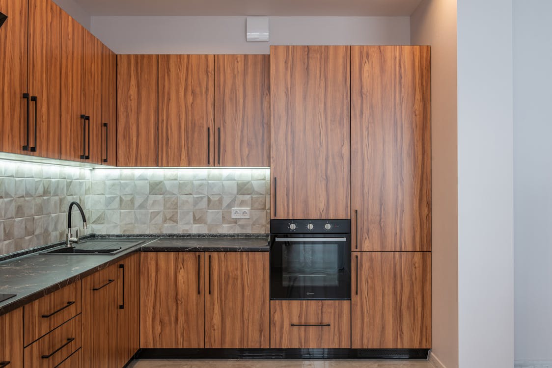 Interior of kitchen with wooden cupboards and contemporary black oven near marble counter with sink and tiled wall in light apartment