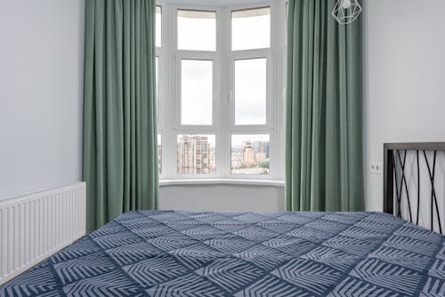 Interior of cozy light bedroom with blue blanket on bed placed against window with green curtain and white walls at home