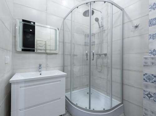 Interior of contemporary light washroom with modern glass shower cabin placed near wall with white sink and mirror at home