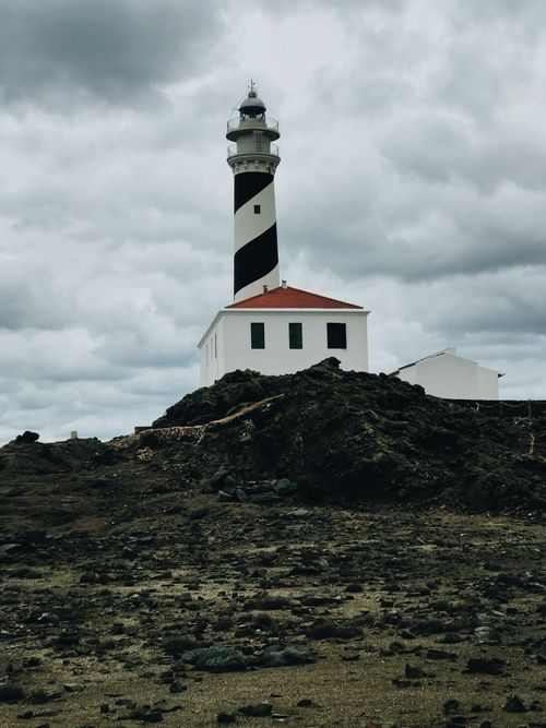 Free Tall lighthouse located near structure on grassy terrain covered with green plants against overcast sky on coastal terrain in nature Stock Photo