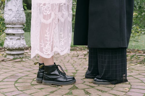Person in White Lace Dress and Black Boots Standing Face To Face With Person In Black Blazer and Trousers