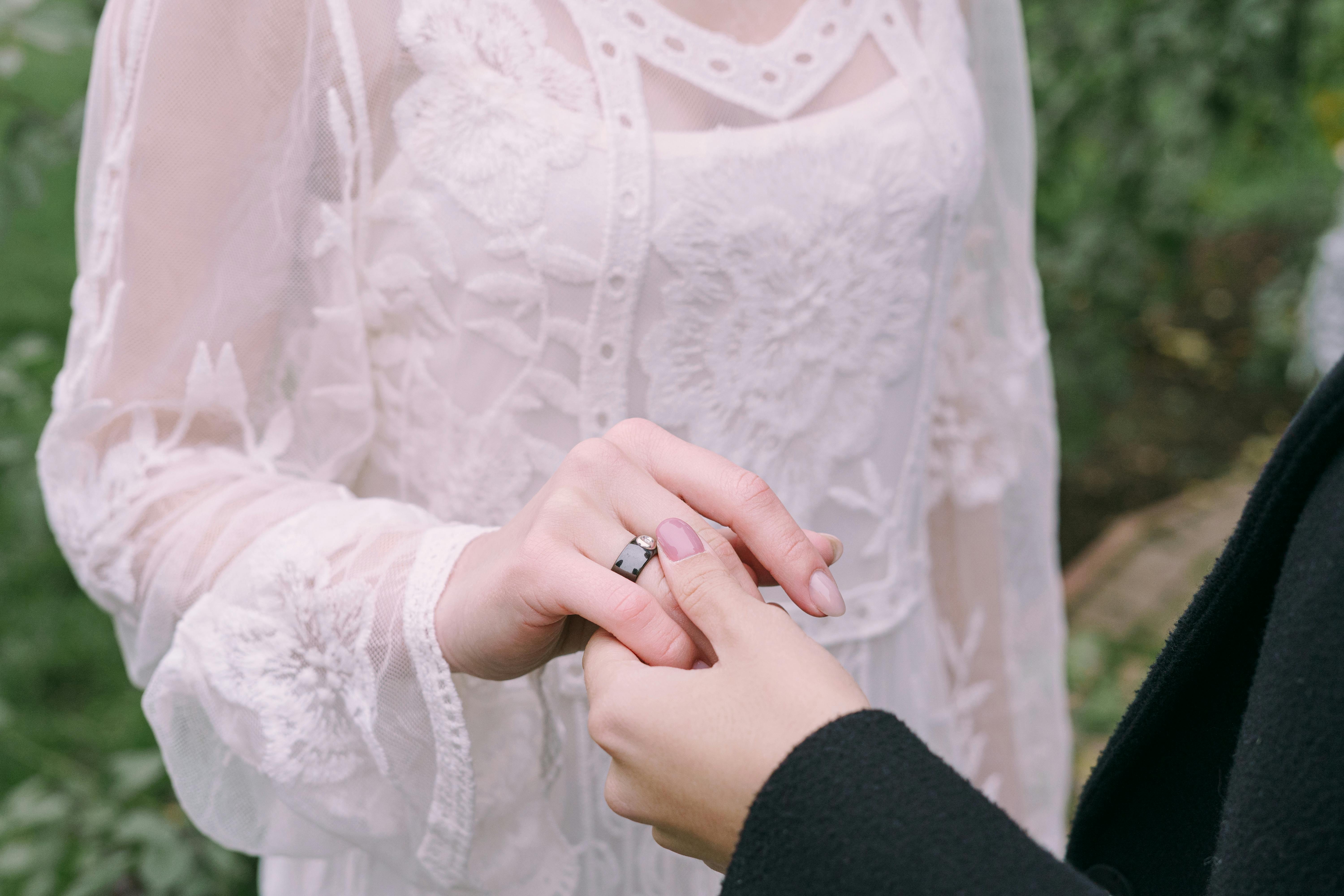 womans hand in black long sleeve holding hand of woman in white laced dress with black ring