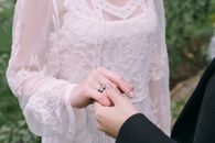 Womans Hand in Black Long Sleeve Holding Hand of Woman in White Laced Dress with Black Ring