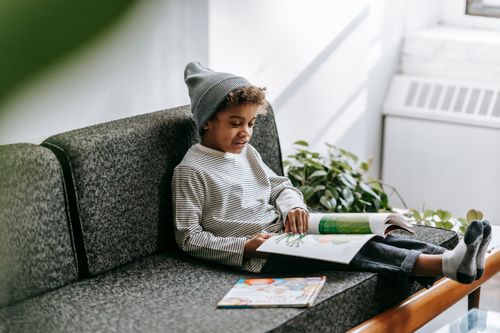 Free Adorable African American clever boy in gray hat and casual outfit reading interesting magazine on sofa of lounge Stock Photo