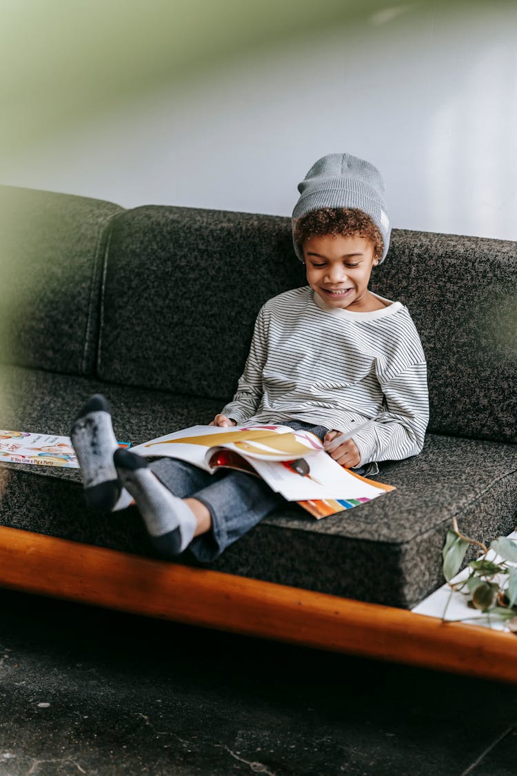 Happy Adorable Black Boy Reading Magazine On Sofa