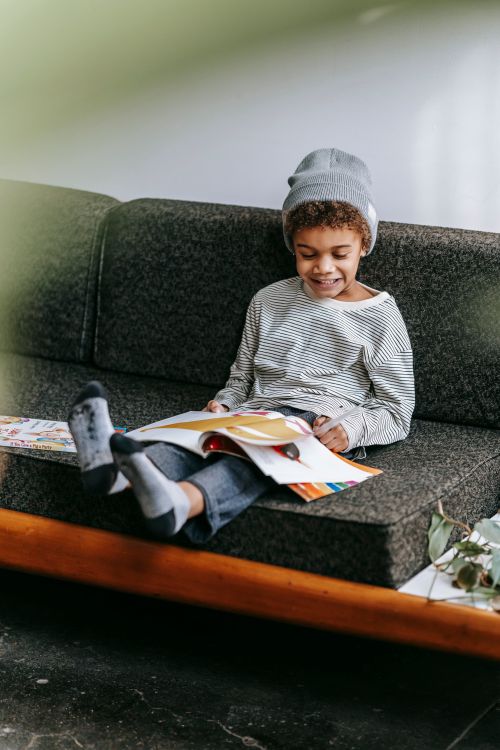 Free Full body of cheerful cute African American boy smiling and reading fascinating magazine on gray sofa of lounge Stock Photo