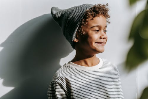 Cheerful African American boy in gray hat looking away and smiling near wall in daytime