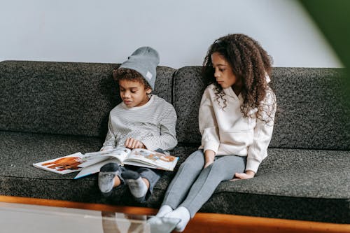 Full body of adorable clever concentrated African American boy and girl reading interesting book on sofa