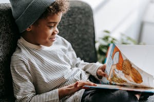 Side view of crop glad African American boy with curly hair smiling and watching pictures of book on blurred background
