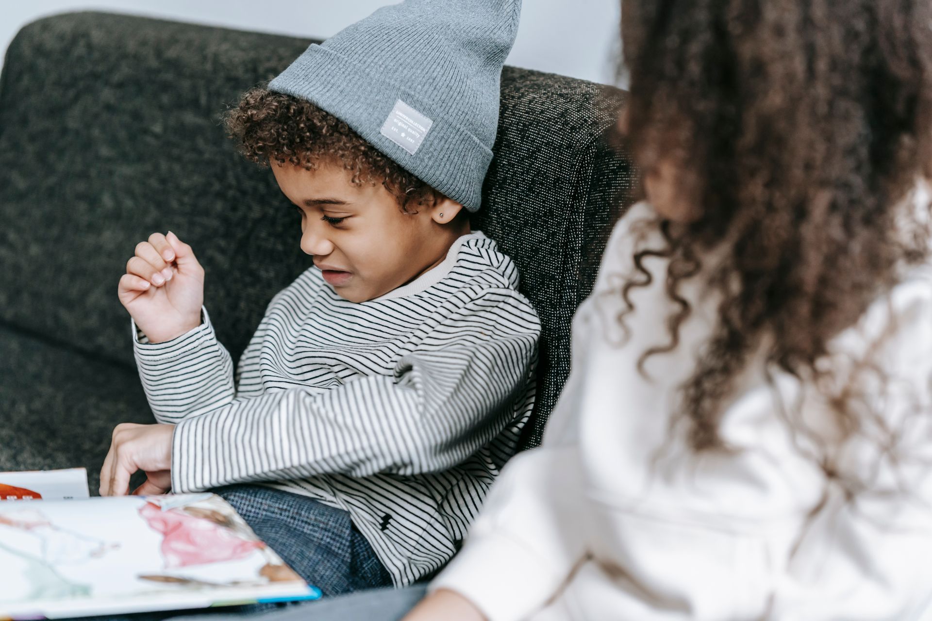 Clever adorable African American boy watching pictures of interesting fairytale in book while crop anonymous sister reading