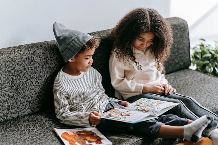 Clever African American brother and sister in trendy outfits reading interesting story with pictures in book on sofa