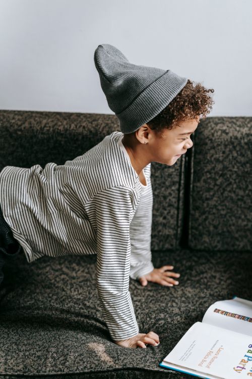 Free Side view of joyful African American boy in gray hat and stylish outfit with interesting book on couch Stock Photo