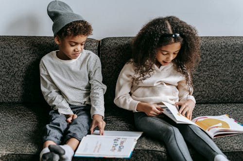 Free Grimacing stylish African American siblings reading books and sitting together on cozy couch in living room Stock Photo