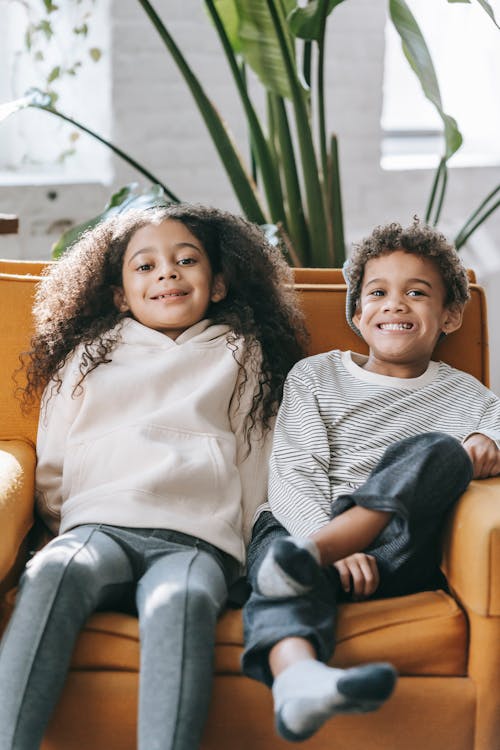 Cheerful stylish African American siblings sitting together on comfy armchair in light living room and looking at camera with toothy smiles