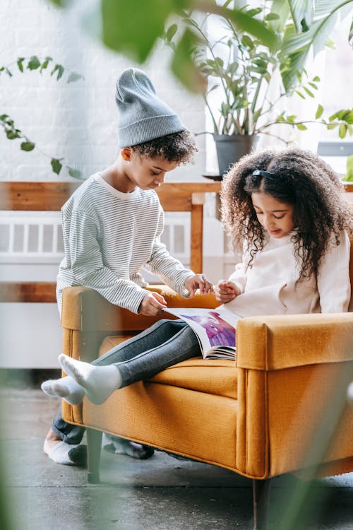Free Cute little black boy in trendy clothes helping sister reading book while sitting on armchair in modern apartment Stock Photo