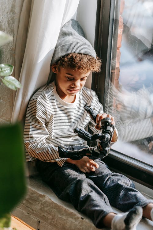 Curious black boy sitting near window and playing with toy at home