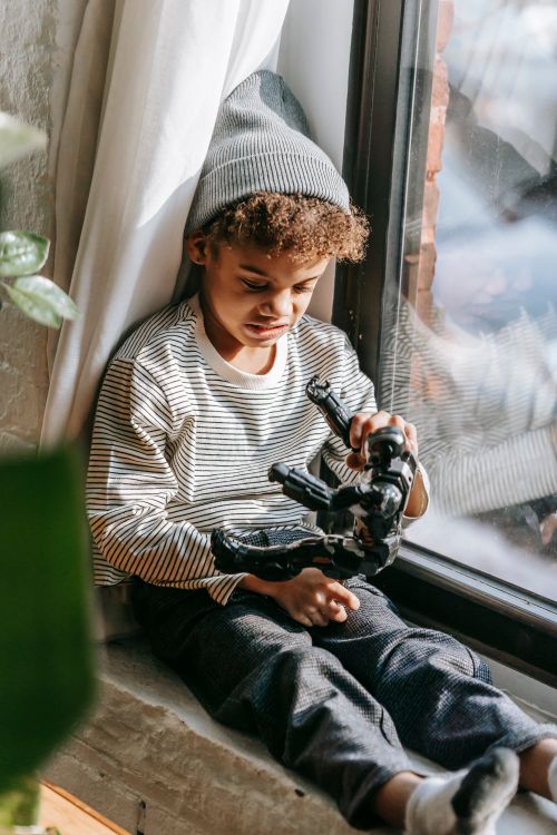 Free Concentrated African American boy in trendy clothes and hat sitting on windowsill and playing with toy in apartment in daytime Stock Photo