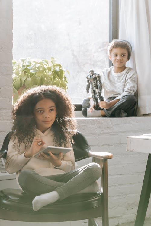 Positive African American girl with long curly hair sitting on chair and browsing tablet while spending time with brother sitting on windowsill and playing with toy at home in daytime