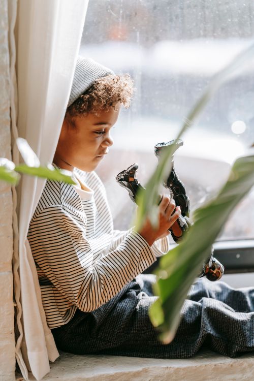 Free Side view of pensive African American boy in casual clothes sitting on windowsill and playing with toy while spending time at home in sunny day Stock Photo