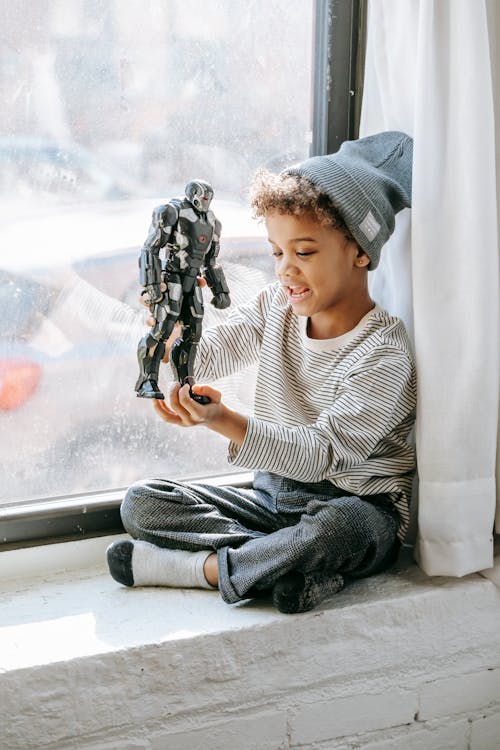 Cheerful African American boy with curly hair in casual clothes and hat sitting on windowsill and playing with toy at home in sunny day