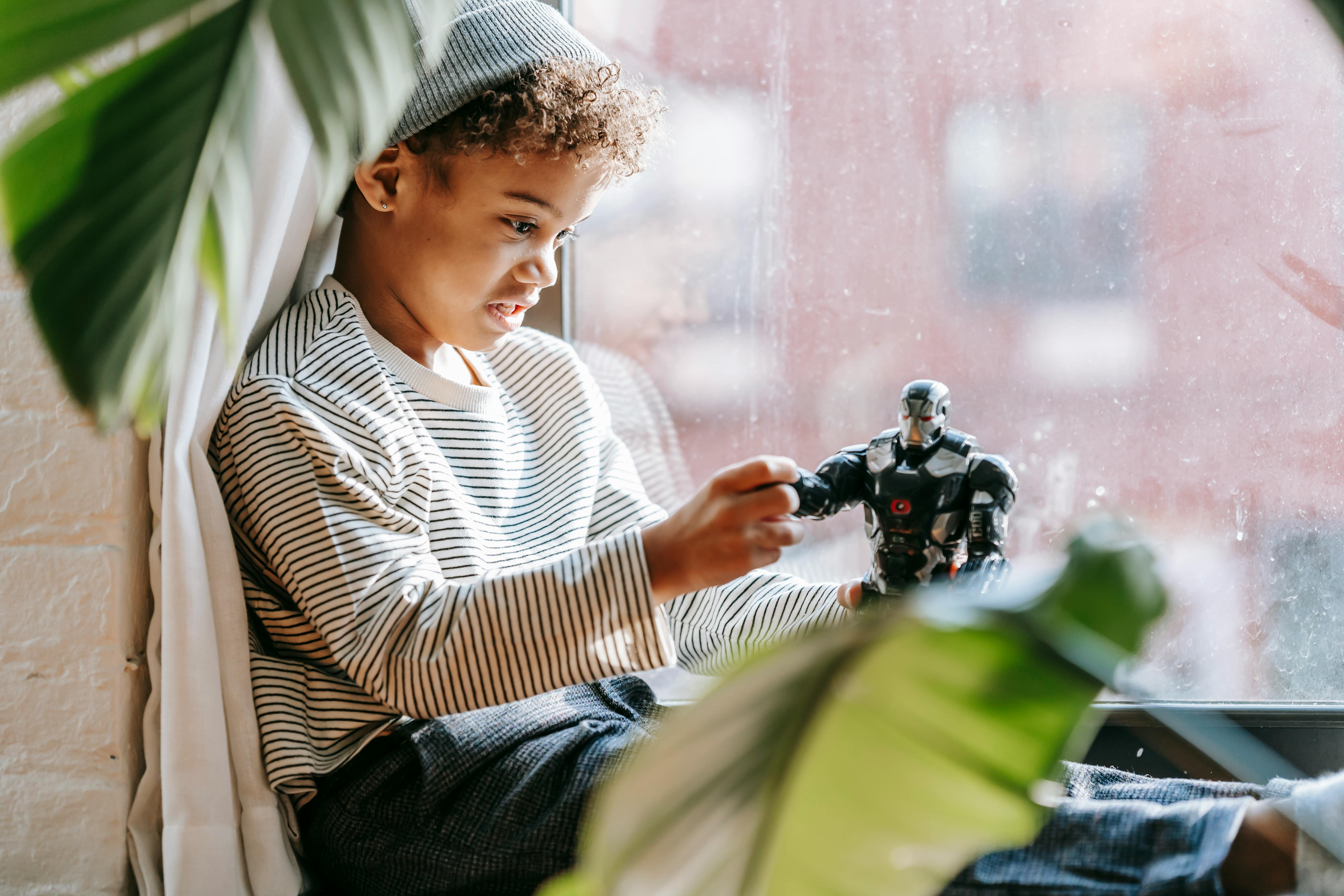 pensive black boy sitting on windowsill and playing with toy