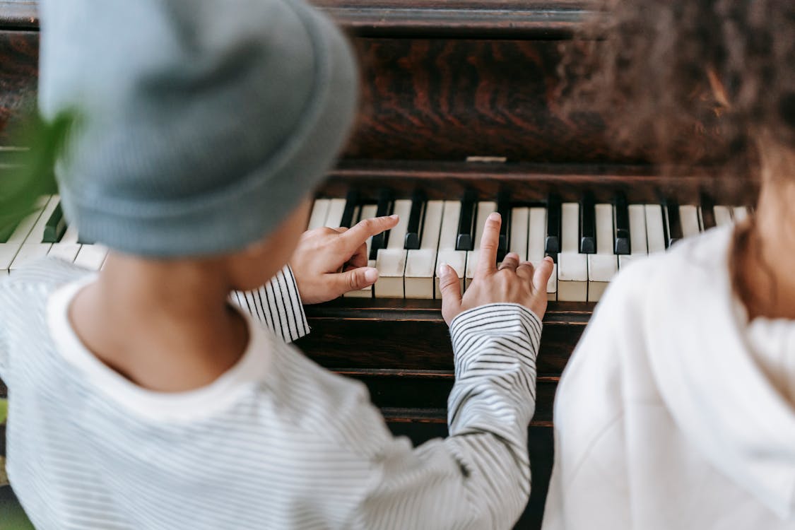 Free Back view of crop anonymous ethnic kids in casual clothes playing piano together in bright room Stock Photo