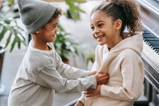 Side view of positive black boy tickling girl belly at bright home wearing casual outfits near piano and potted plants with green leaves