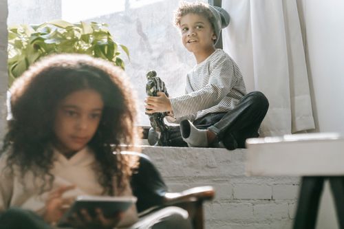 Free African American girl sitting in armchair with tablet near brother sitting on windowsill and playing with robot toy near plant with green leaves and curtains near window in light apartment Stock Photo