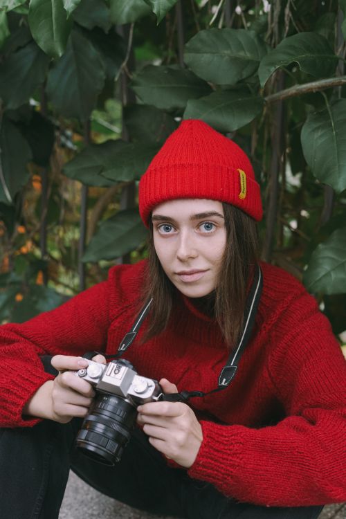 Free Portrait of a woman in red beanie and sweater holding a camera in a lush green environment. Stock Photo