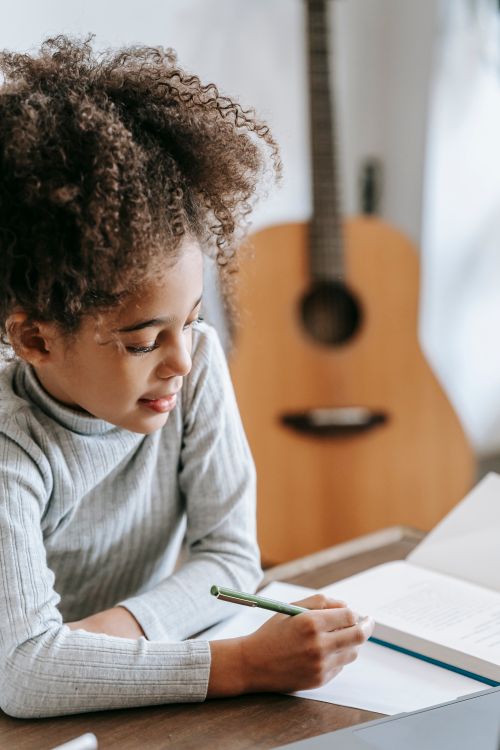 Free Side view of positive African American girl with curly hair writing on paper while sitting at table with book while studying at home with guitar on blurred background Stock Photo