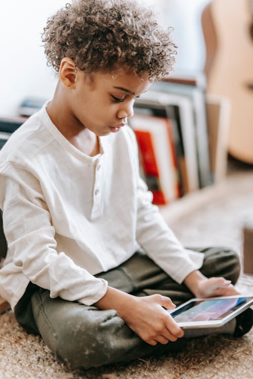 Full body side view of concentrated Africa American boy playing video game on tablet while sitting with crossed legs near books in room with guitar on blurred background