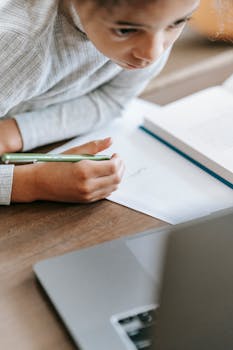 A young girl intently studies and takes notes with a laptop, book, and paper.