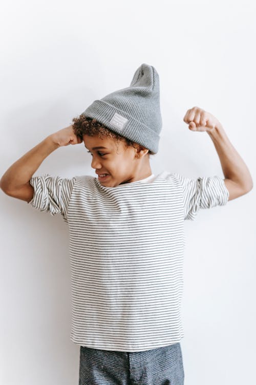 Free Positive African American boy in trendy hat and striped outfit showing strength while standing against white background in light room Stock Photo