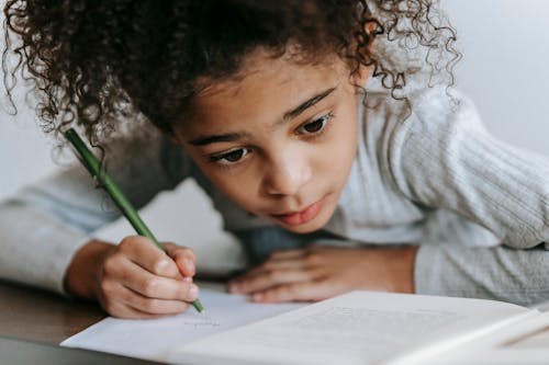 Crop concentrated African American girl with curly hair sitting at table with textbook while taking notes on sheet of paper
