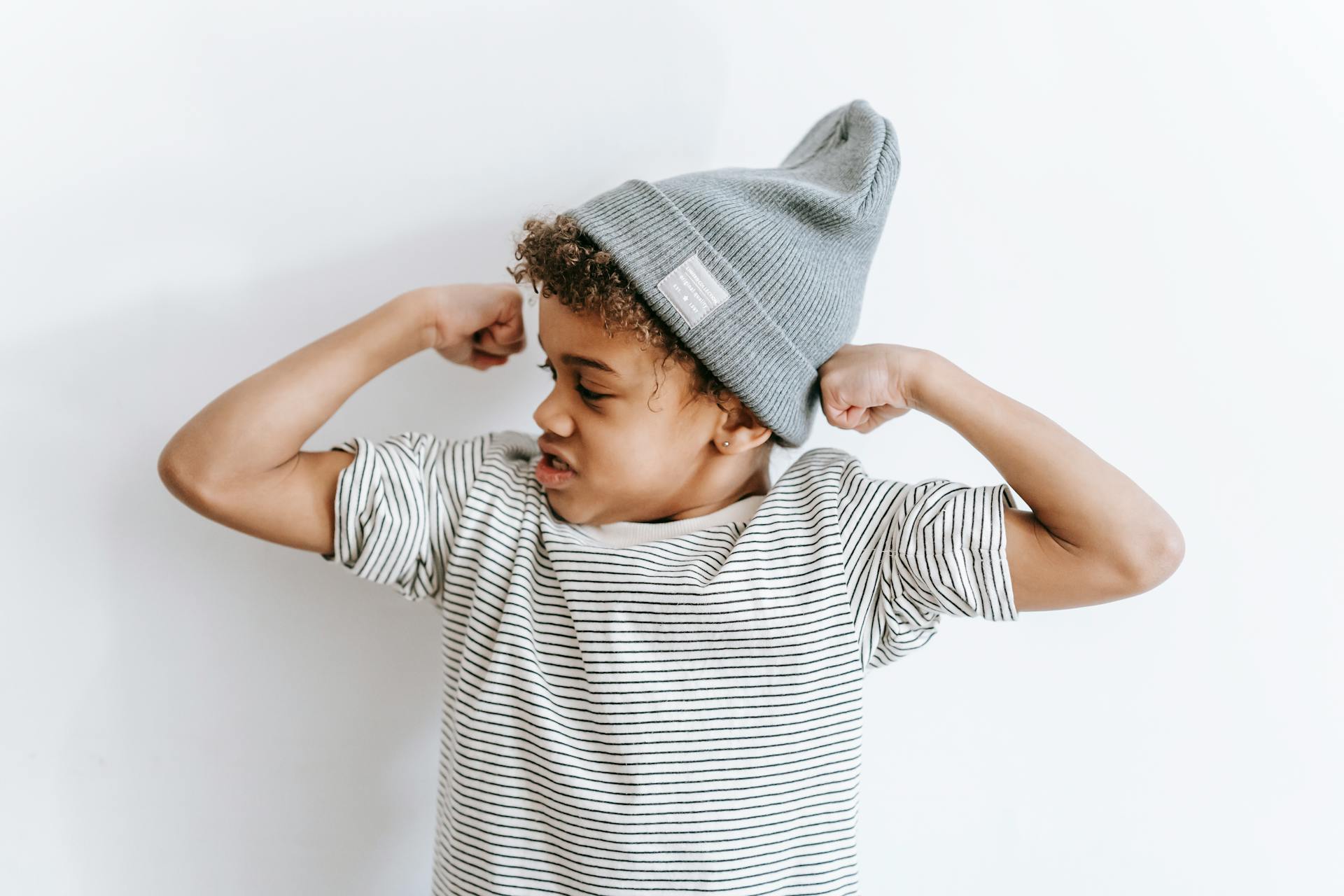 Strong African American boy in stylish hat showing biceps while standing on white background with arms bent in elbows in light room