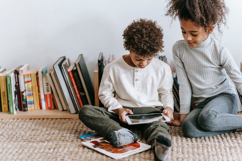 Concentrated African American boy browsing tablet while sitting on floor with black sister in room with abundance of books at home