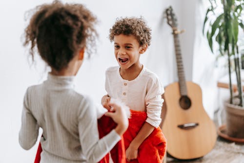 Free Funny African American boy looking at anonymous sister while standing with Santa Claus costumes in room with guitar during Christmas holiday Stock Photo