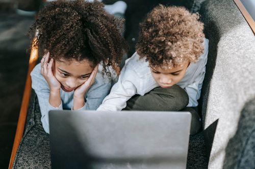 Free From above of concentrated African American siblings browsing modern netbook while lying on comfortable couch in living room at home Stock Photo