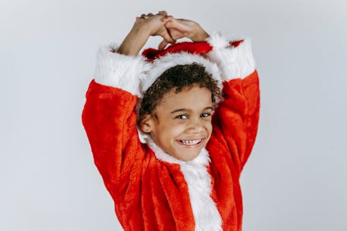 African American child in Santa costume with toothy smile and raised arms looking at camera on light background