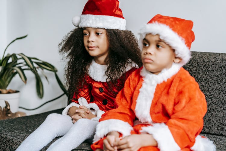 Black Siblings In Santa Costumes Resting On Sofa At Home