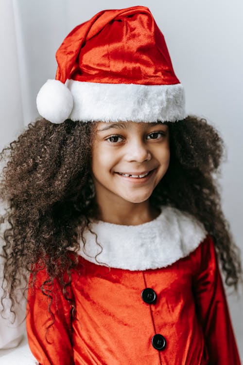 Cheerful African American kid with curly hair in Santa costume looking at camera on Christmas day on white background