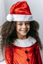 Cheerful African American kid with curly hair in Santa costume looking at camera on Christmas day on white background