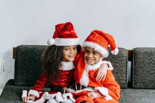 Content black siblings in Santa costumes embracing on couch during New Year holiday and looking away in house
